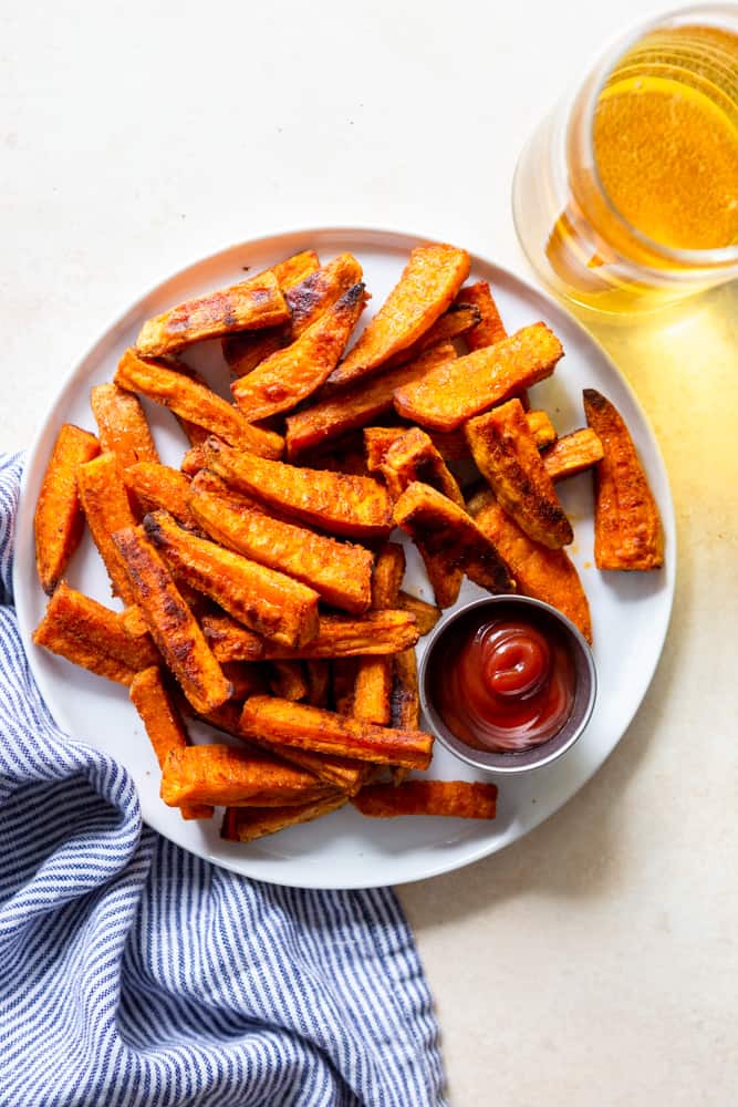 A plate of  crispy oven baked sweet potato fries with a side of ketchup and a glass of beer.