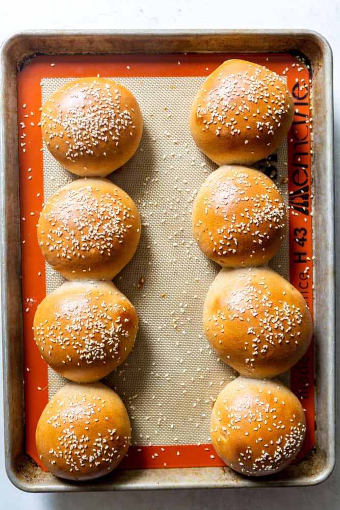 A baking sheet lined with silpat holding 8 golden hamburger buns topped with sesame seeds