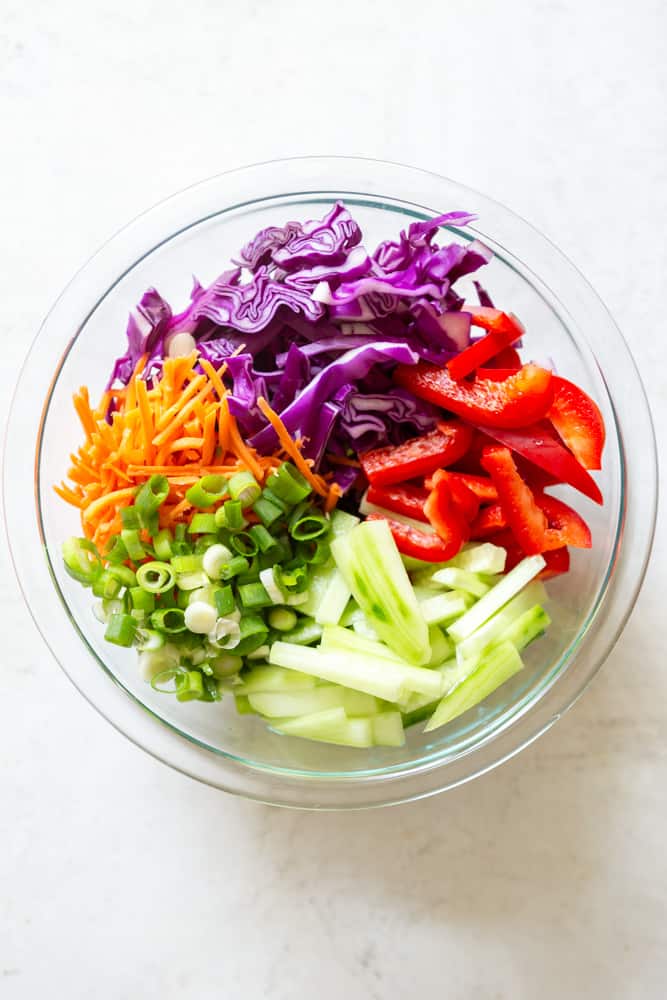 Vegetables for cold peanut noodle salad in a glass bowl. Grated carrots, purple cabbage, red bell pepper, cucumber matchsticks, sliced green onion.