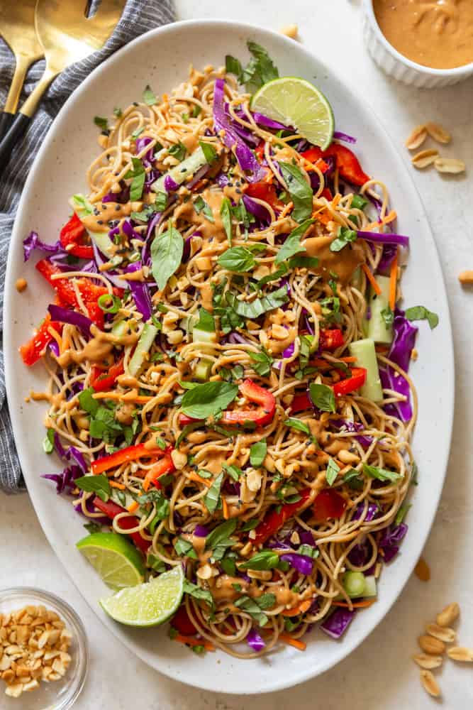 Cold peanut noodle salad on a large white platter.  Garnished with peanut sauce, basil, chopped peanuts and sesame seeds.