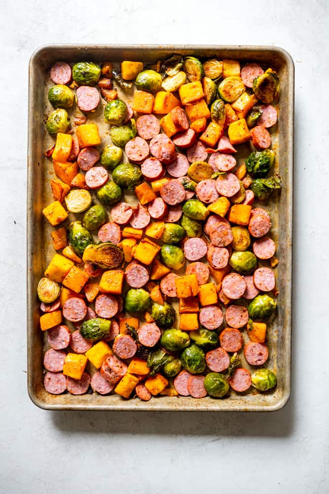 Sheet pan with partially cooked brussels sprouts and butter nut squash, with sliced sausage links just added.