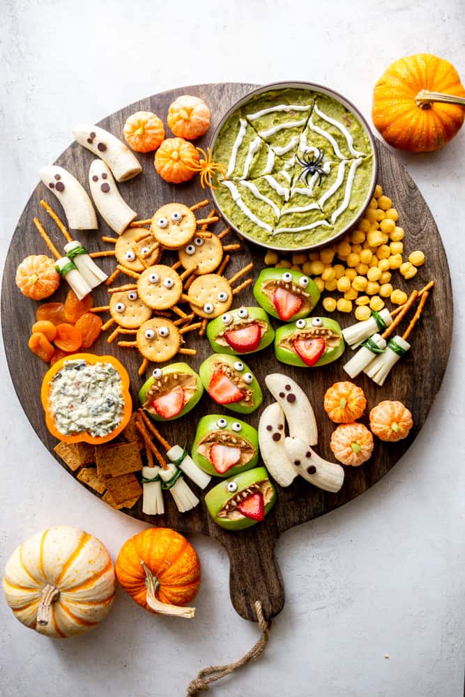 A large wooden charcuterie board with Halloween themed snacks from this post - boo-nanas, clementine pumpkins, apple monsters, spider crackers, witches brooms, spider web guacamole.  Also some crackers, cheese puffs, dried apricots and spinach dip in a hollowed out orange bell pepper. There are pumpkins next to the board.