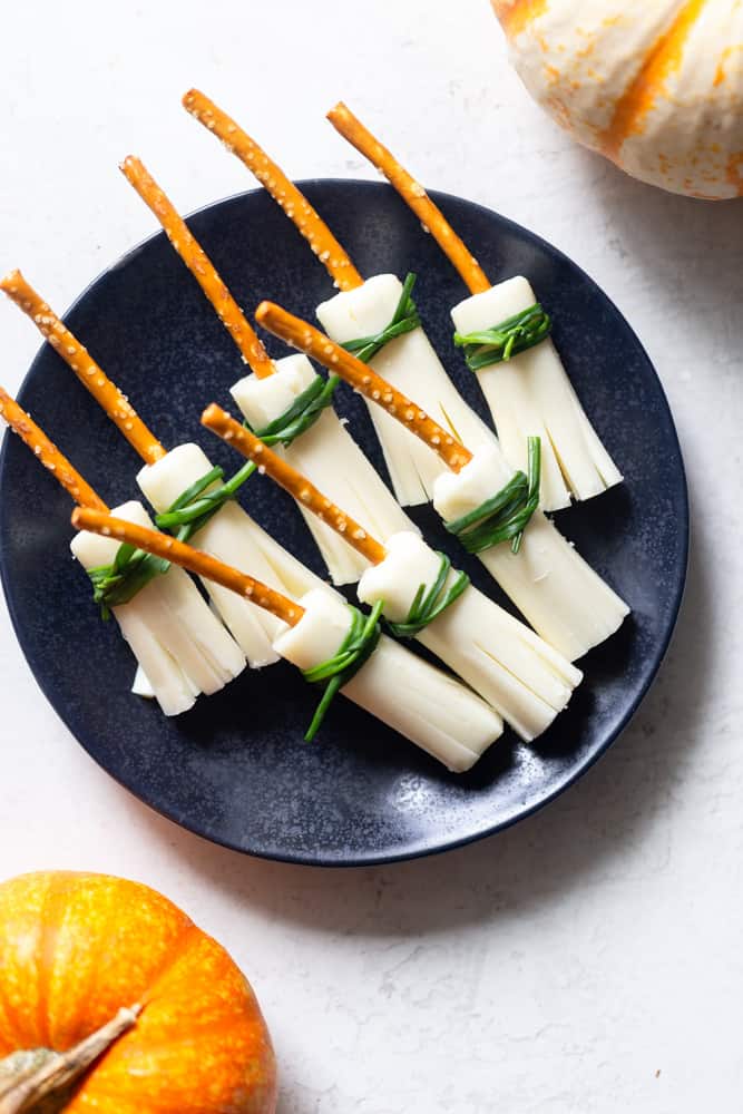 Witches Broom snacks for Halloween on a black plate. Witches brooms are made from string cheese for the bristles, pretzel sticks for the broom handles and chives tied around the tops of the cheese to finish the look.