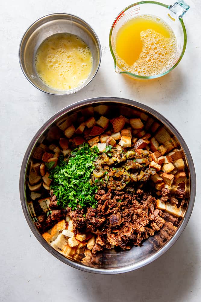Large steel bowl with bread cubes, sausage, parsley and sauteed vegetables. A glass measuring cup of chicken broth, a small bowl of beaten eggs.