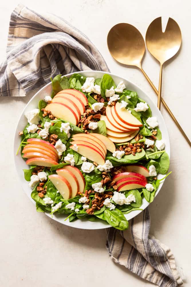 Spinach apple salad with goats cheese and chopped candied pecans in a large white bowl.