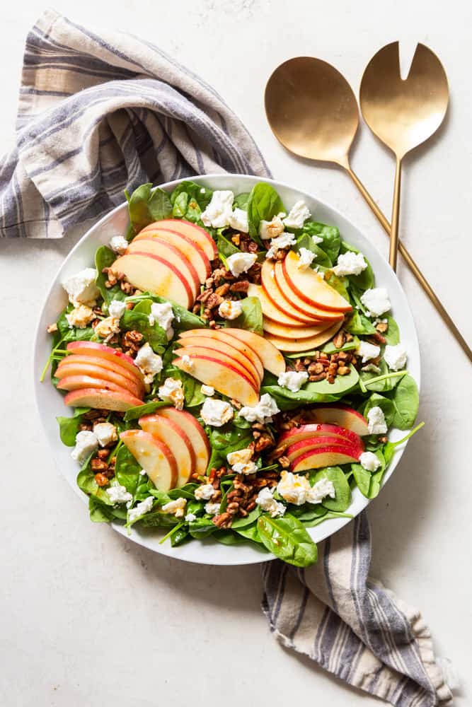 Spinach apple salad with goats cheese and chopped candied pecans in a large white bowl, drizzled with apple cider vinaigrette. 