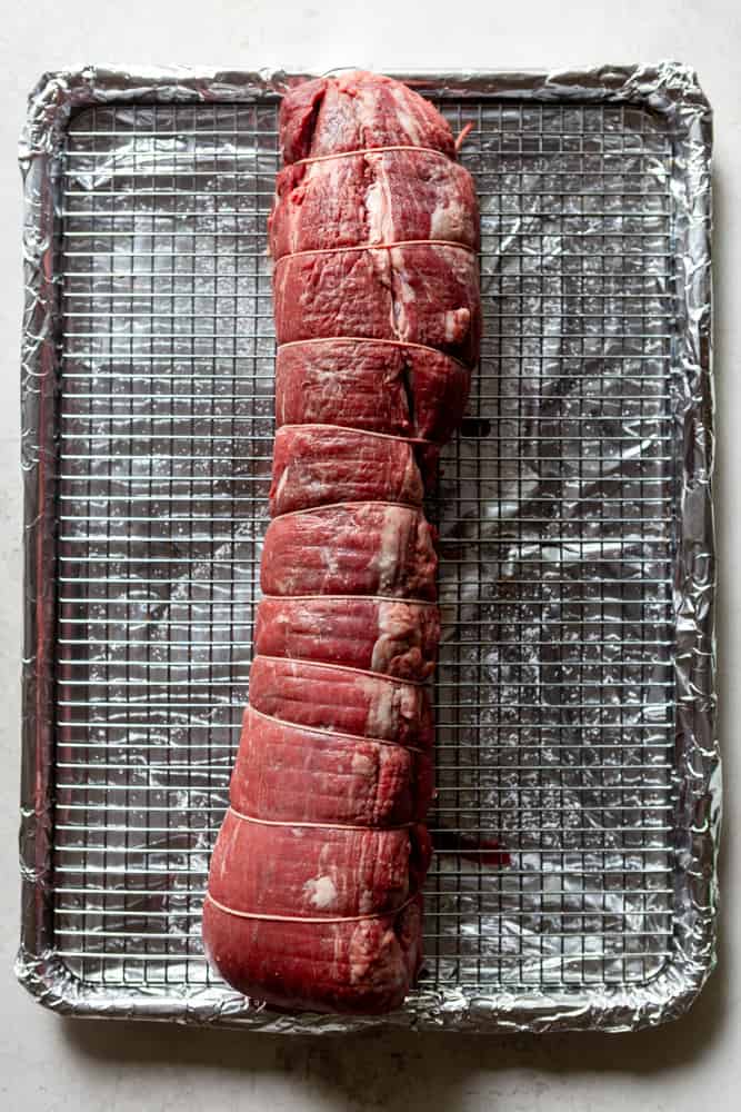 Uncooked Beef Tenderloin ties with twine and sitting on a cooling rack over a sheet pan lined with foil.