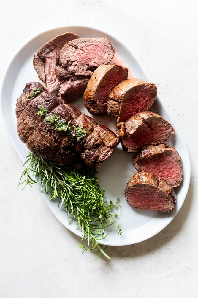 Slow Roasted Beef tenderloin garnished with fresh herbs on a white platter.