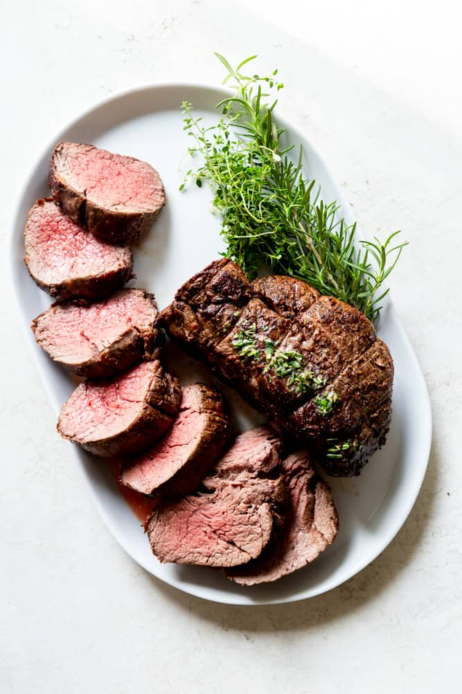 A beef tenderloin roast and some fresh green herbs on a white platter.