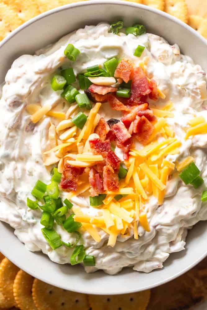 Close up of Million Dollar Dip in a white bowl sitting on a wooden board, surrounded by buttery round crackers. Garnished with Bacon, shredded cheese, slivered almonds and green onions. 