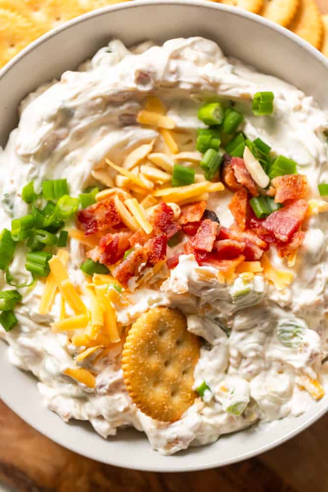 Million Dollar Dip garnished with crispy bacon pieces, grated cheddar cheese, green onions, slivered almonds.  A buttery cracker is being dipped. 