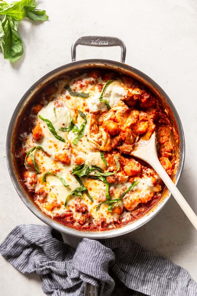 large stainless steel skillet with a red marinara sauce, gnocchi and sausage, topped with melted cheese and garnished with basil.