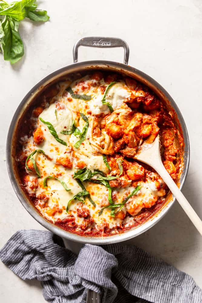  large stainless steel skillet with a red marinara sauce, gnocchi and sausage, topped with melted cheese and garnished with basil.