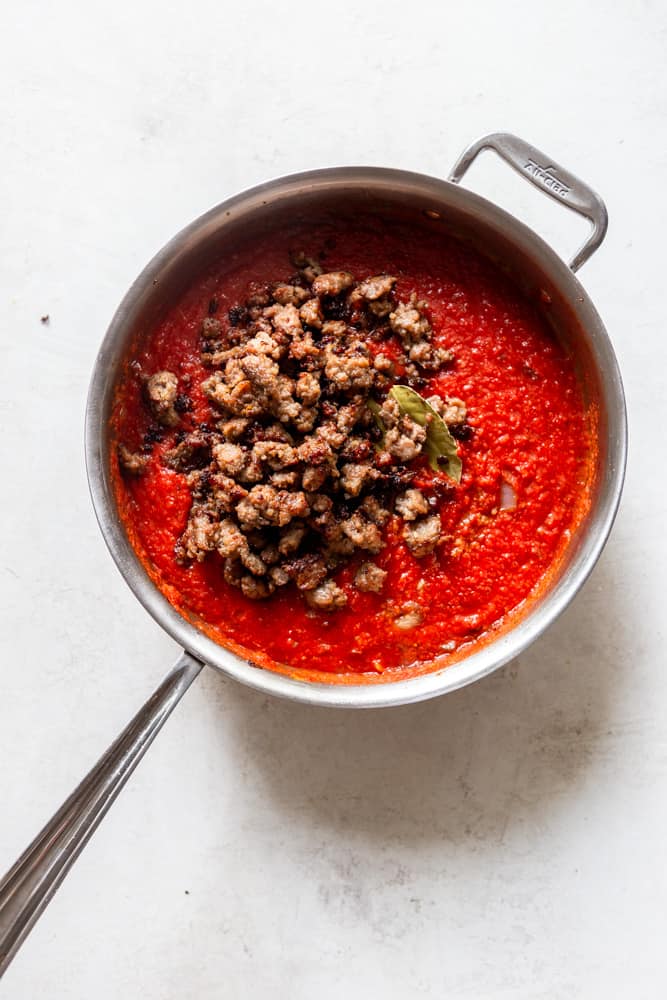 A skillet with tomato marinara, topped with browned ground sausage and a bay leaf