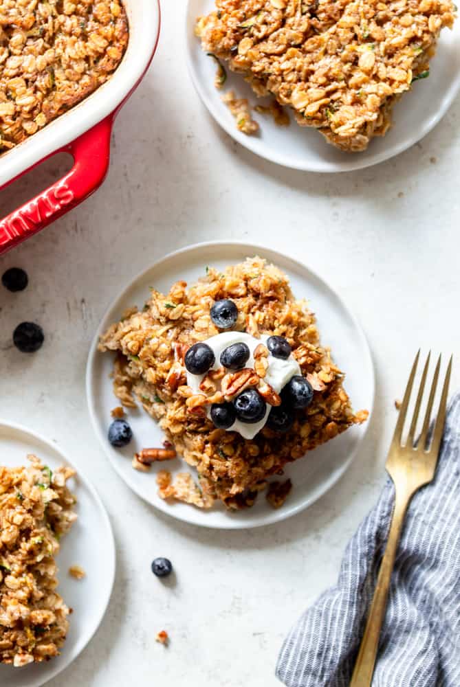 A slice of Zucchini Baked Oatmeal on a white plate, topped with a dollop of yogurt, blueberries and pecans.