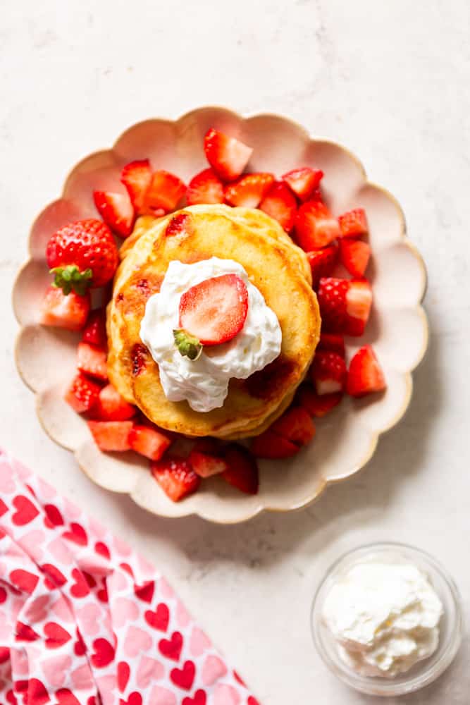 Strawberry pancakes topped with whipped cream and a fresh strawberry, on a scalloped plate, next to a bowl of whipped cream and a napkin with pink and red hearts.
