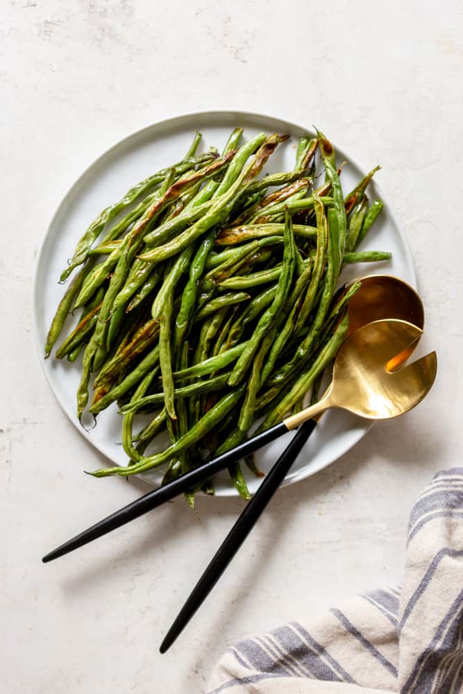 Roasted Green Beans on a white plate with fold and black utensils. On a white background, with a grey and blue striped dish towel nearby.
