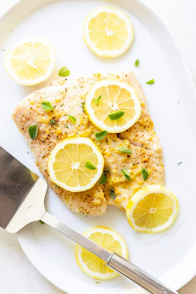 Garlic Butter Baked Halibut Filet on a shite platter, garnished with lemon slices and fresh oregano. 