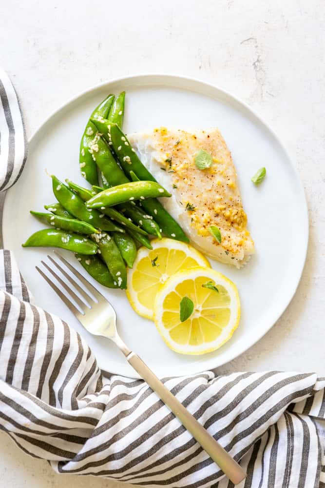 Garlic Butter Baked Halibut on a white plate with snap peas and lemon slices.