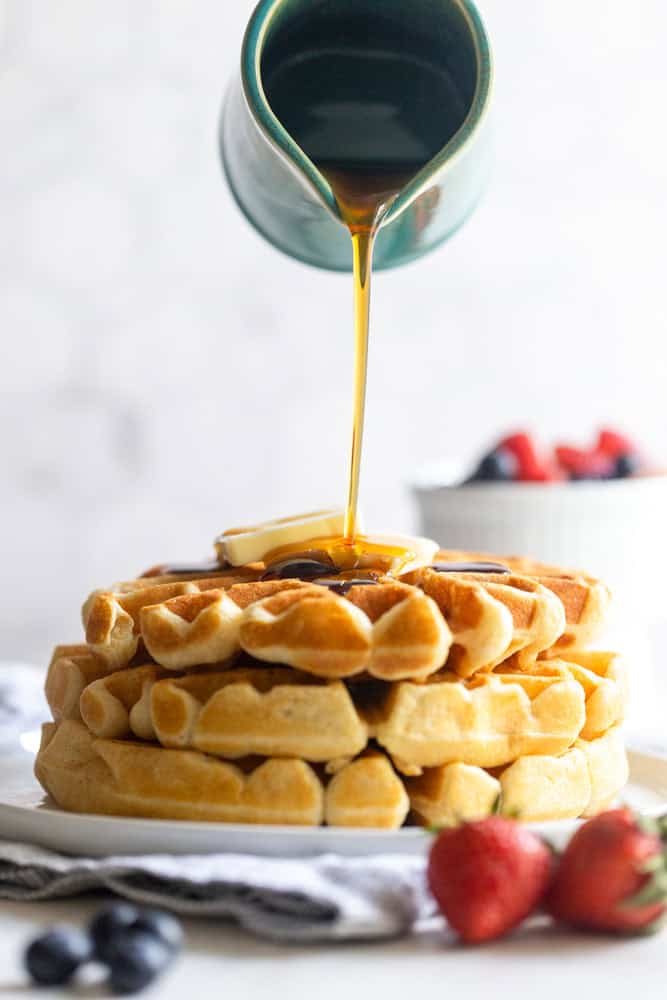A stack of fluffy buttermilk waffles with maple syrup being poured over them. 
