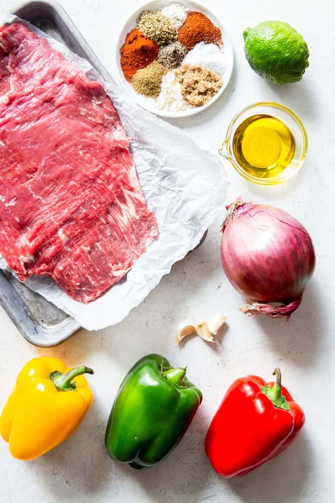 Ingredients for steak fajitas - flank steak, spices, lime, olive oil, red onion, garlic, yellow, green and red bell peppers.