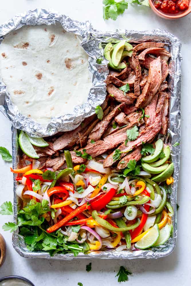 Sliced flank steak, onions and bell peppers on a sheet pan along with foil wrapped tortillas.