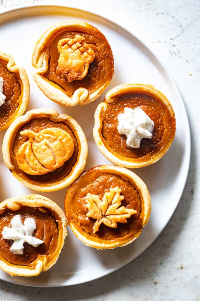 mini pumpkin pies on a white plate, topped with whipped cream and decorative pie crust cookies. 